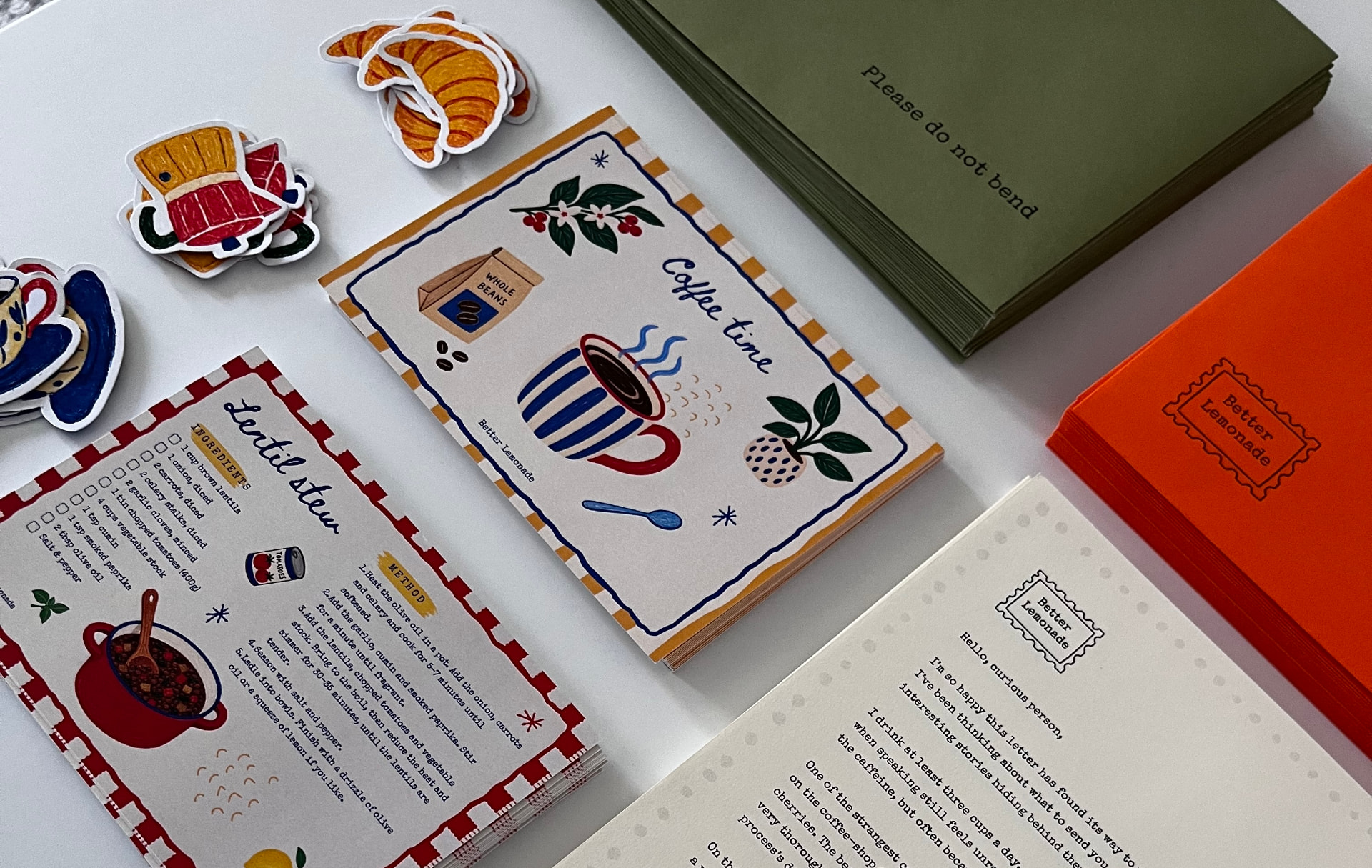 Olive and orange envelopes, illustrated stickers, a recipe card and a typewritten page laid out on a white table